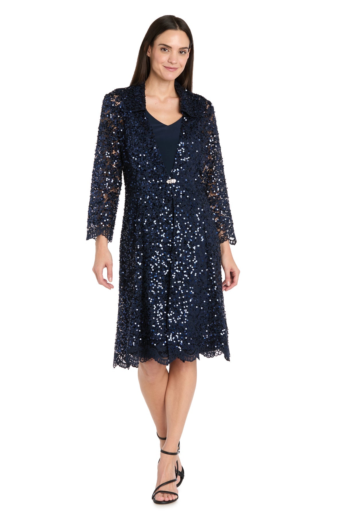 A woman wearing the R&M Richards 3742 Short Navy Jacket Party Dress with long sleeves and black sandals stands against a white background.