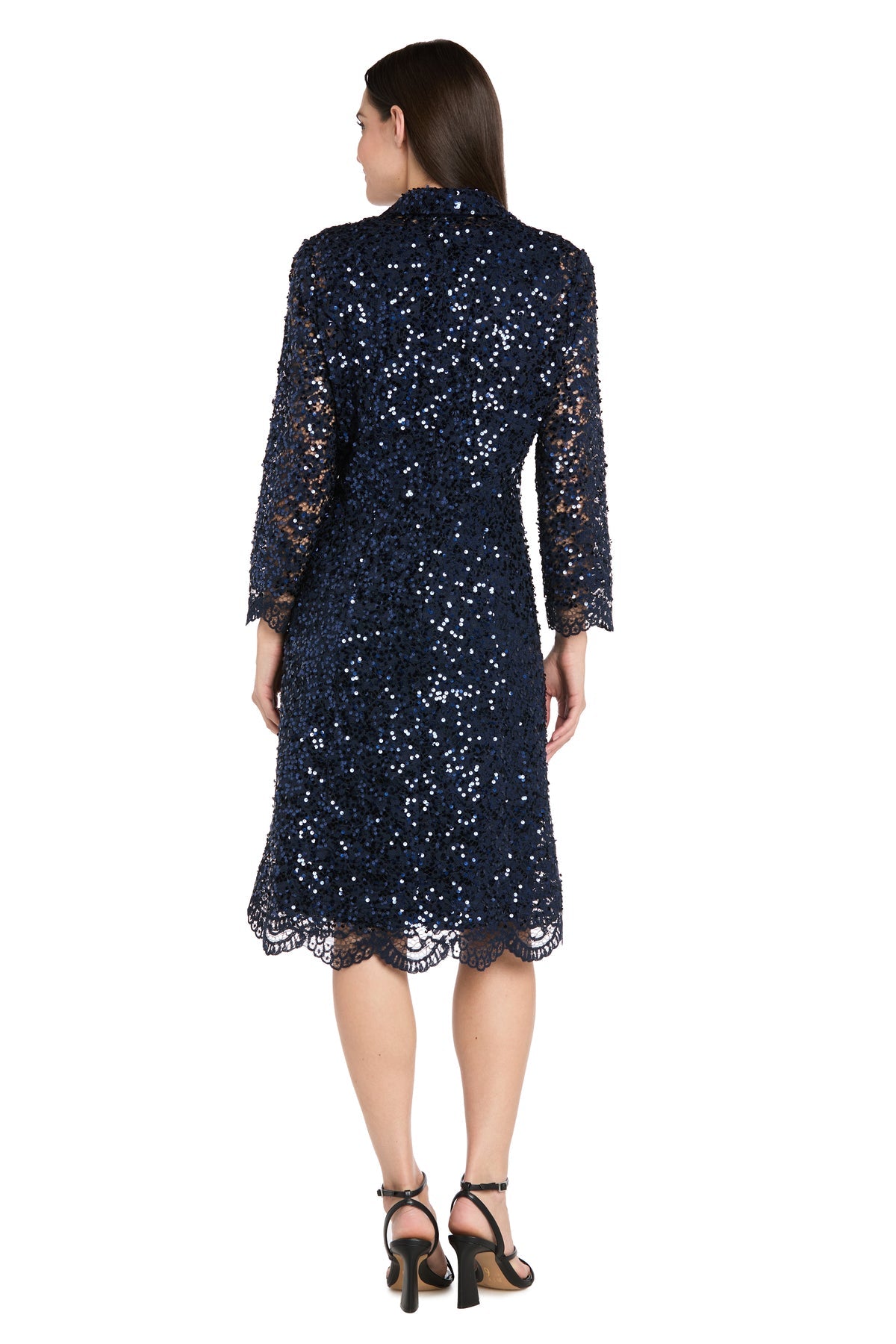 A woman stands with her back turned, wearing the Short Navy Jacket Party Dress: R&M Richards 3742, a knee-length dress with long sleeves and scalloped hem, paired with black heeled sandals.