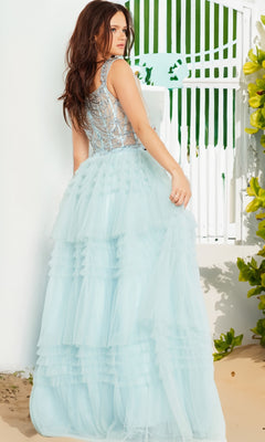 A woman in the Jovani 37438 Sheer-Corset Ruffled Prom Ball Gown, featuring a light blue sleeveless A-line silhouette and beaded corset bodice, stands outdoors by a white wall and gate amid greenery.