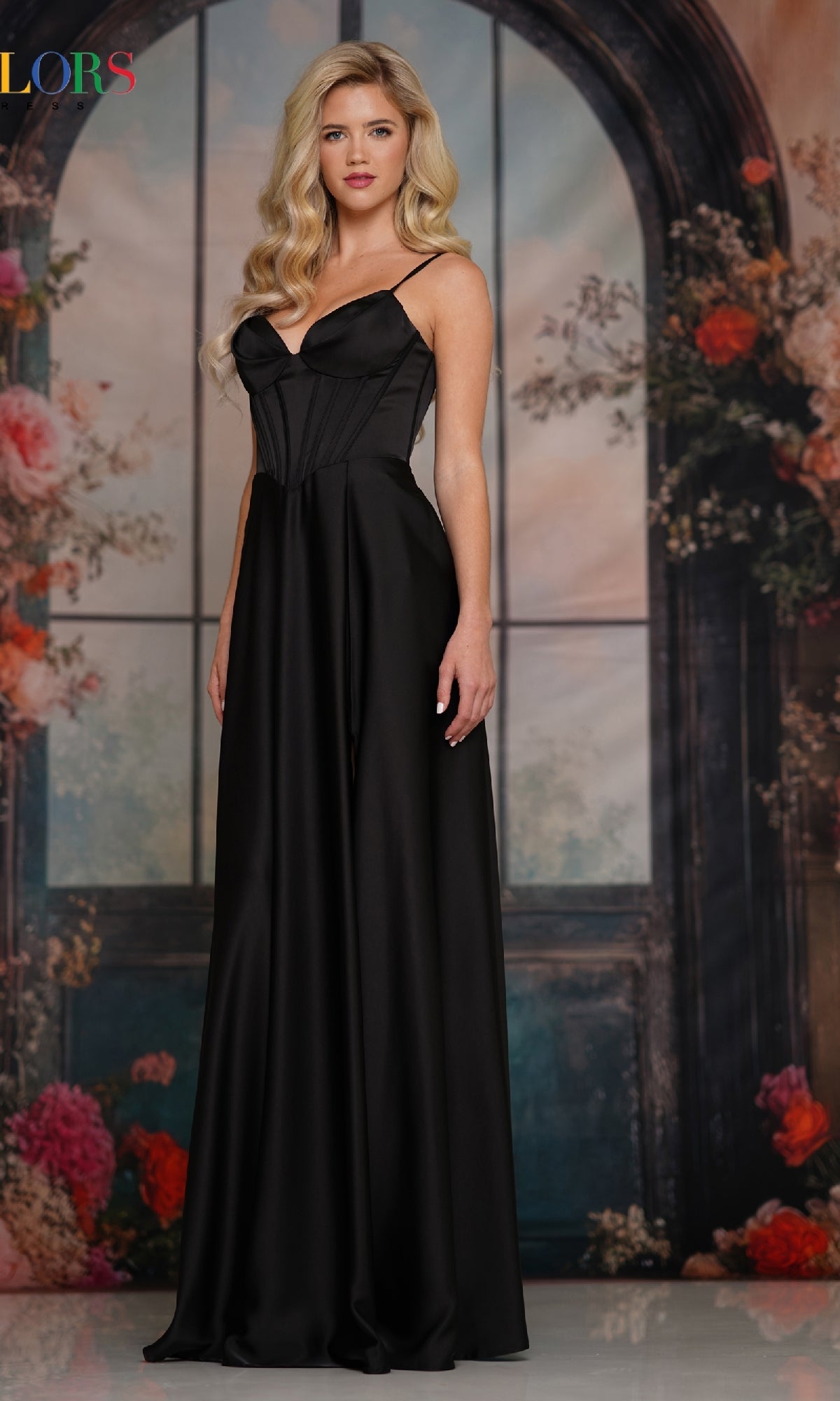 A woman in the Colors Dress 3743, a long satin corset prom dress, stands before a floral backdrop with a large arched window, her elegant evening gown shimmering in the light.