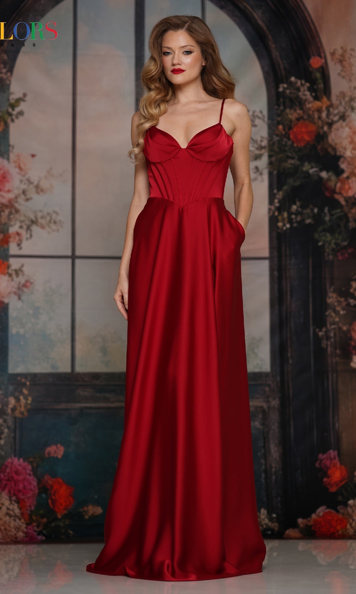 A woman in the Colors Dress 3743, a long wine satin corset prom dress, stands before a floral background with an arched window design.