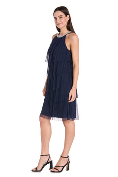 A woman in profile models the Petite Short Navy Party Dress: R&M Richards 3744P, featuring a tiered design and paired with black heeled sandals, set against a white background.