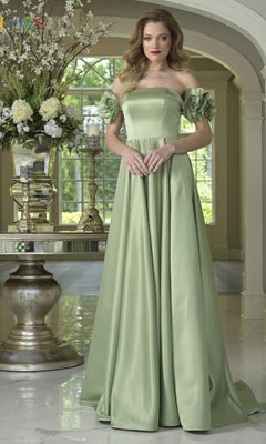 A woman wears the Colors Dress Off-Shoulder Satin Prom Gown (Style 3744) with floral sleeves, standing indoors by a table with vases and white flowers, framed by large arched windows.