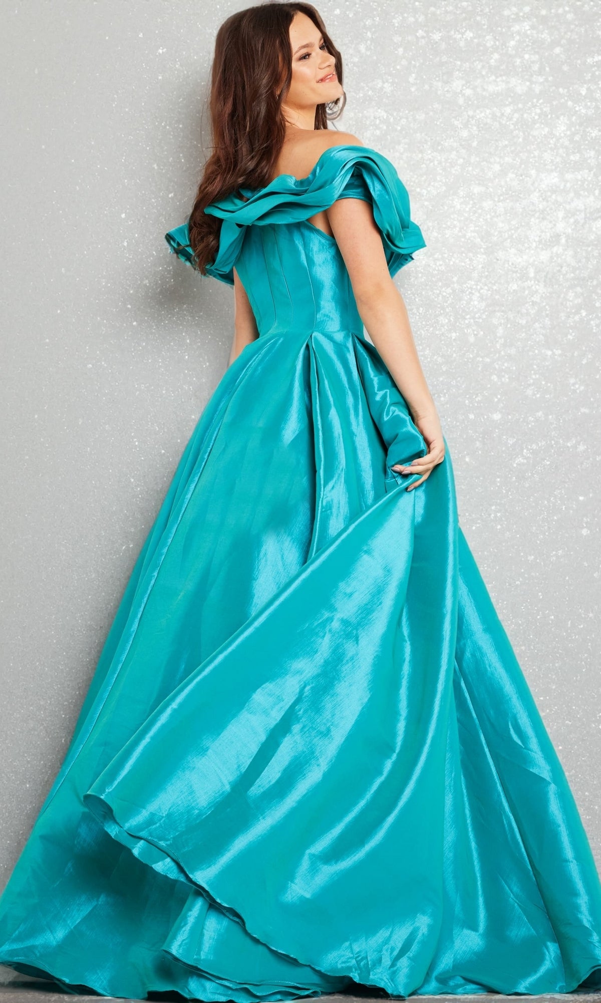 A woman wearing the Long Prom Dress 37476 by Jovani, featuring a teal hue and off-the-shoulder ruffled sleeves, poses against a glittery silver backdrop while holding her dress.