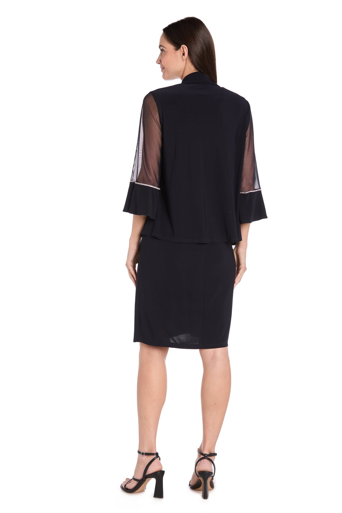 A woman stands with her back to the camera, wearing the Petite Two-Piece Party Dress: R&M Richards 3747P—navy blue, knee-length, featuring sheer flared sleeves—and black heeled sandals.