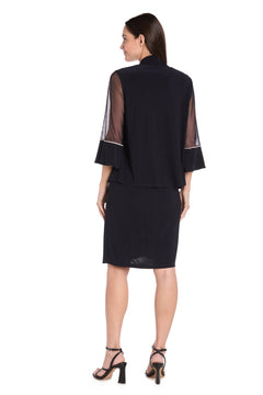 A woman stands with her back to the camera, wearing the Petite Two-Piece Party Dress: R&M Richards 3747P—navy blue, knee-length, featuring sheer flared sleeves—and black heeled sandals.