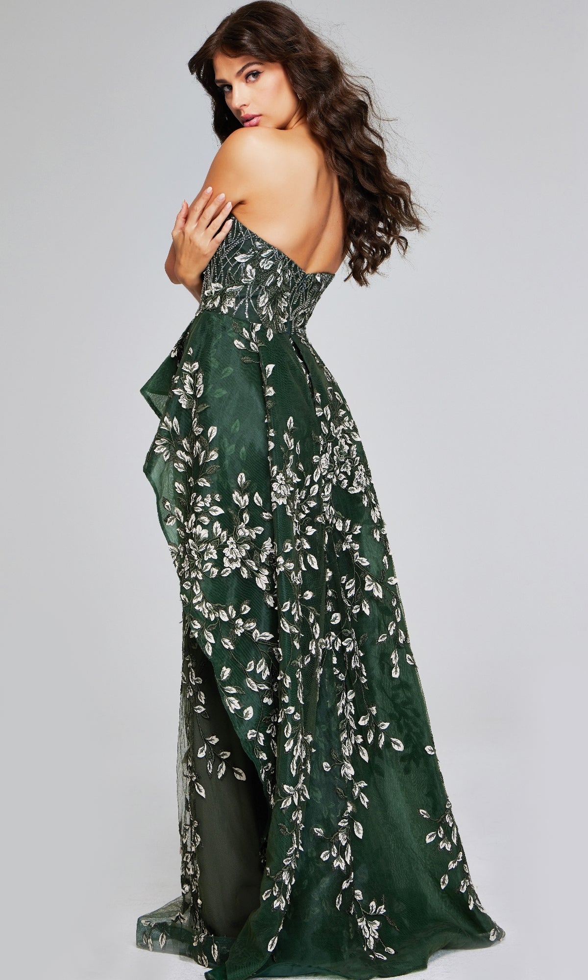 A woman poses in the Long Dark Green Formal Dress: Jovani 37599, a strapless, floor-length gown featuring silver floral embroidery and a low back, set against a plain grey background.