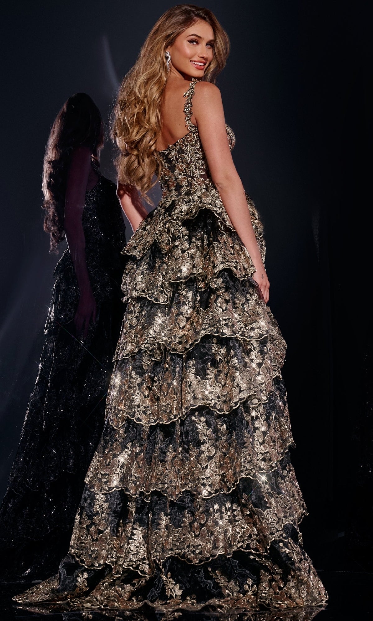 A woman with long wavy hair poses sideways in front of a dark background, wearing the Long Prom Dress 37630 by Jovani—a gold and black floral tiered ball gown—with her reflection visible.