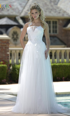 A woman in the Colors Dress 3796, a white halter-neck long prom dress with 3D floral appliques, stands outdoors in front of a building and greenery.