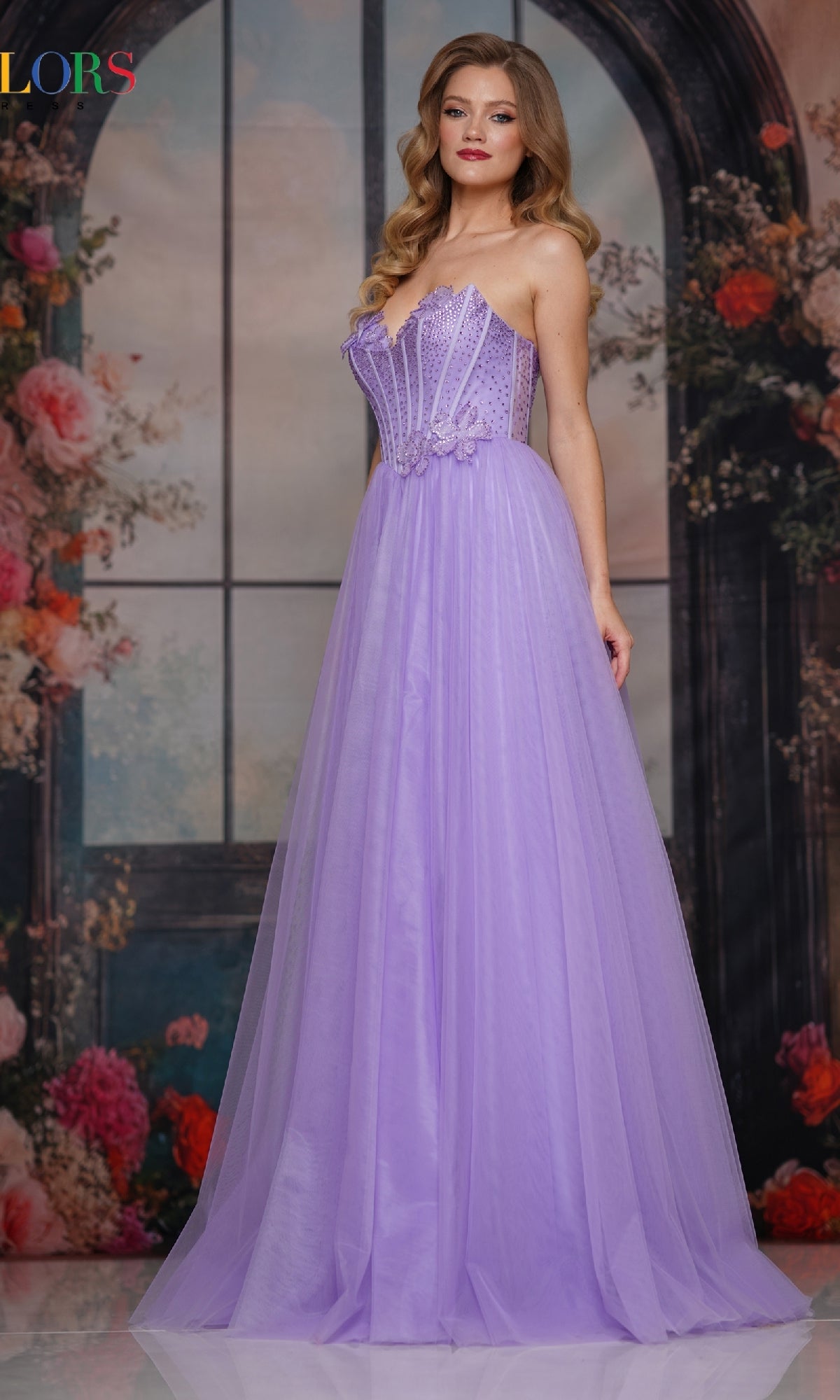 A woman in a lavender Strapless Long Corset Prom Gown (Colors Dress 3797) with 3D floral appliques poses elegantly on a white floor before a floral and window backdrop.