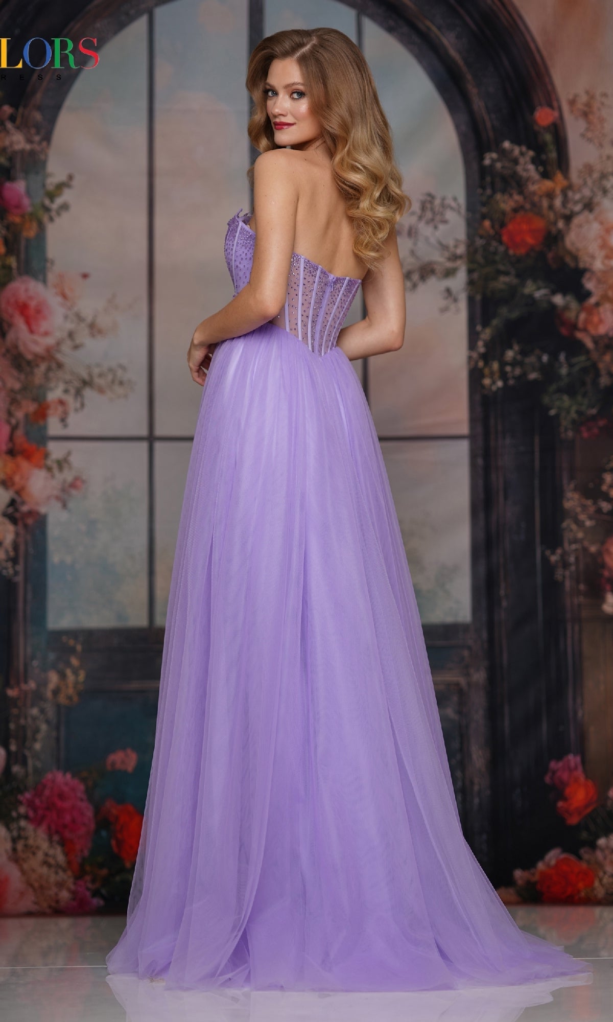 A woman with long wavy hair models the Colors Dress 3797, a strapless long corset prom gown, posing with her back turned before a floral and arched window backdrop.