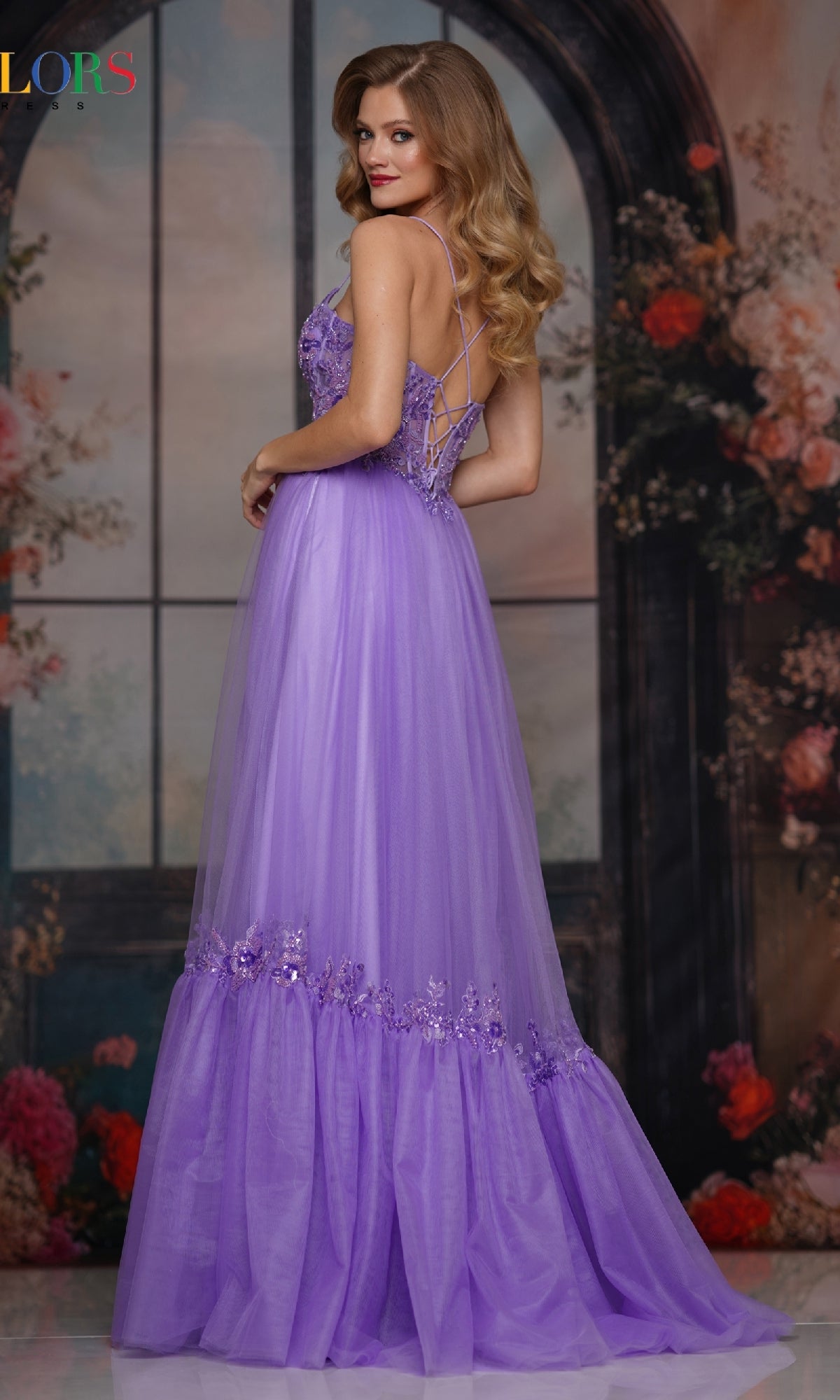 A woman in the Colors Dress 3799, a long lavender prom gown with a sheer embroidered bodice, lace-up open back, and floral embellishments, poses sideways among flowers before a large arched window.