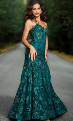 A woman in the Jovani 38004 Long Corset Mermaid Prom Dress, featuring emerald green embroidered lace, stands on an empty tree-lined road, looking at the camera.