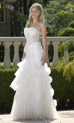 A woman stands outdoors near a stone railing in the Colors Dress 3800 long prom dress, featuring a strapless fitted bodice, lace-up open back, and tiered tulle skirt, smiling amid lush greenery.