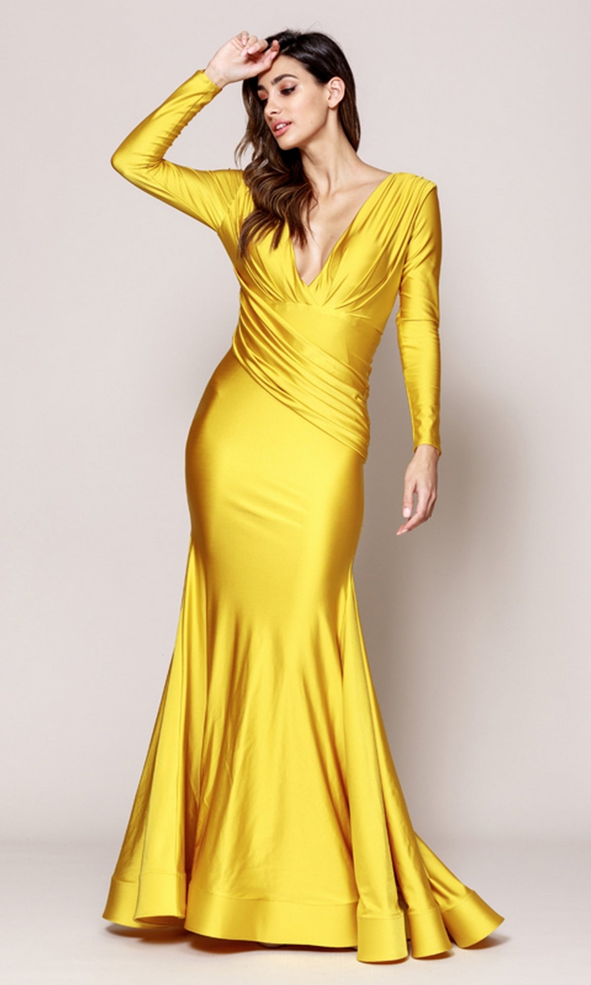 A woman stands against a plain background wearing the Amelia Couture 381: a fitted, long-sleeve, floor-length yellow gown with a deep V-neckline and mermaid skirt.