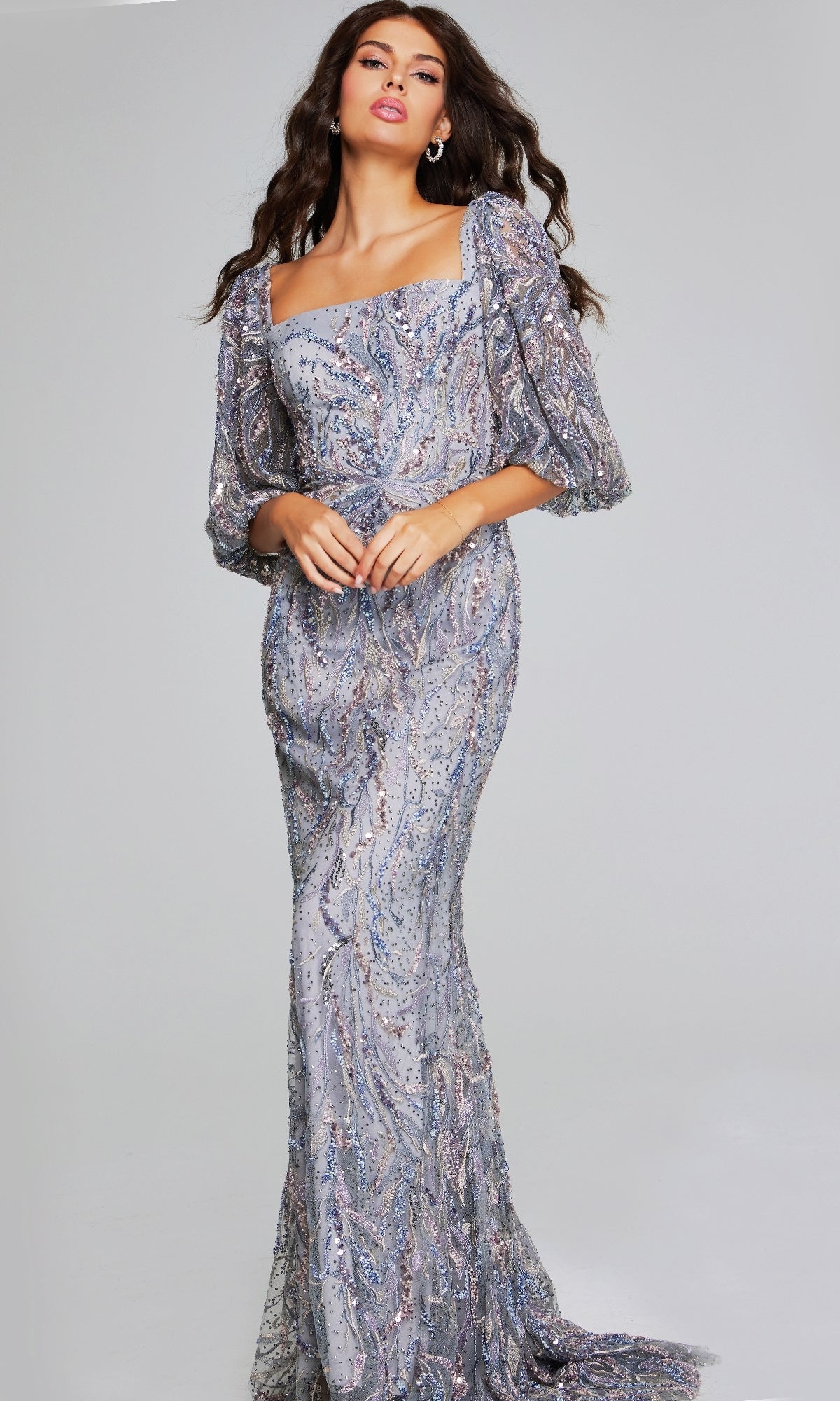 A woman stands against a plain backdrop wearing the Jovani 38110 long grey formal dress, featuring sparkly embellishments and three-quarter sheer sleeves.