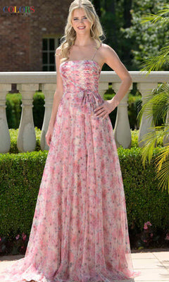 Woman poses outdoors by a stone railing in the Colors Dress 3812—a strappy, long pink prom dress with floral print and bow at waist—surrounded by lush greenery.