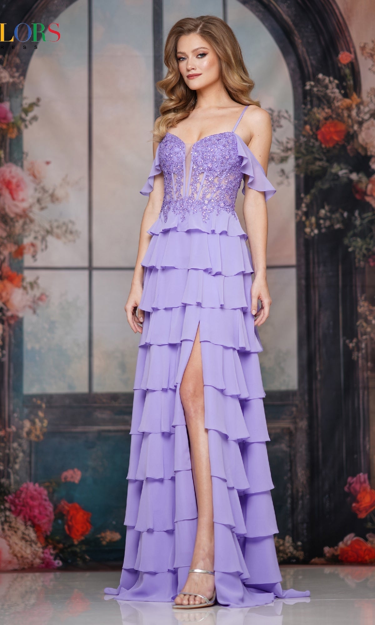 A woman poses in the Colors Dress 3820, a lavender long prom dress with tiered ruffles, spaghetti straps, a corset bodice, and a thigh-high slit, standing before a floral and arched window backdrop.