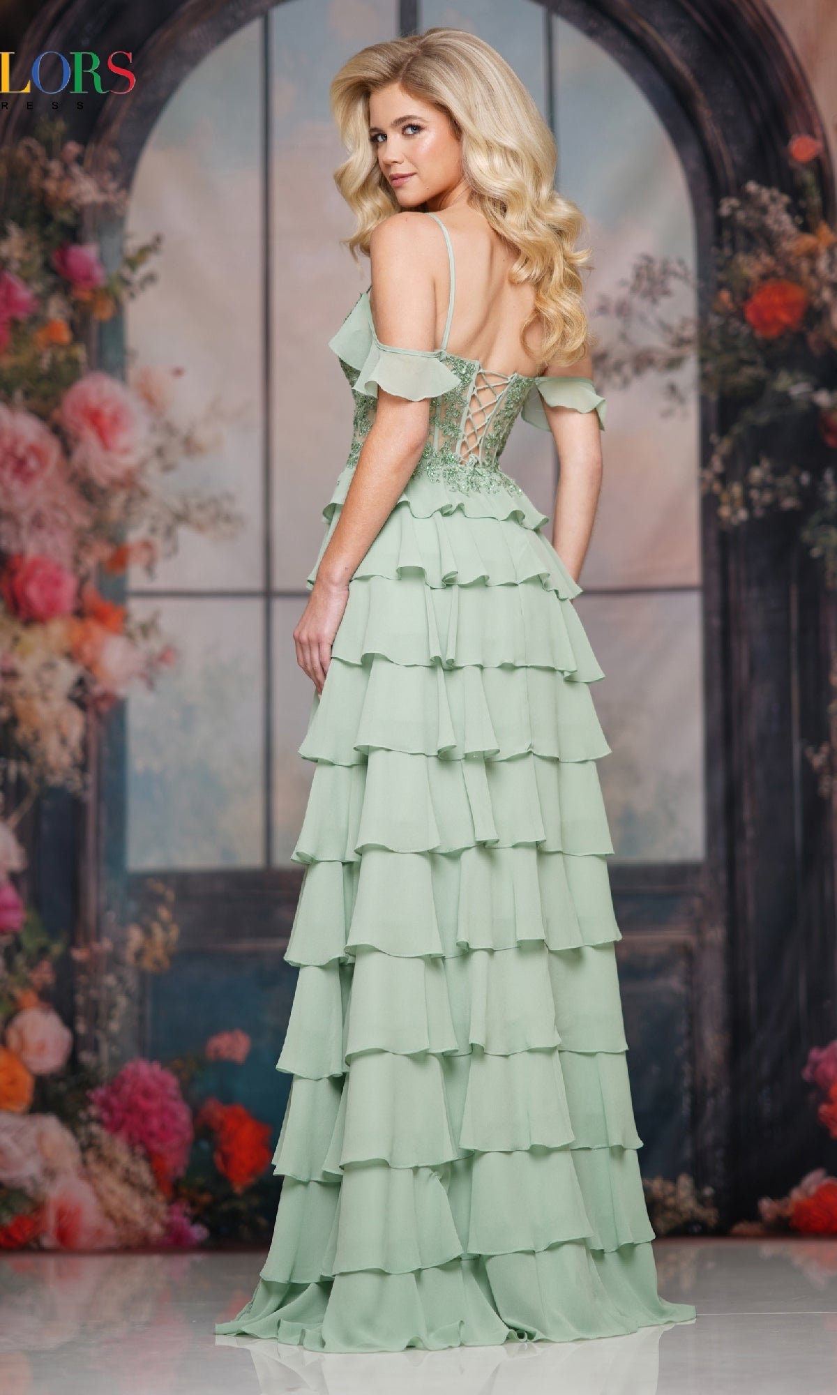 A woman stands facing away before a floral backdrop and arched window, wearing the Colors Dress 3820—a light green, tiered ruffle long prom dress with a lace-up back.