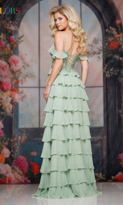 A woman stands facing away before a floral backdrop and arched window, wearing the Colors Dress 3820—a light green, tiered ruffle long prom dress with a lace-up back.