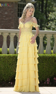 A woman stands outdoors wearing the Colors Dress 3820, a long yellow prom dress with lace, spaghetti straps, a corset bodice, and tiered off-the-shoulder design. She looks down with one hand on her hip.