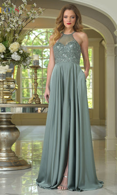 A woman wears the Colors Dress High-Neck Halter Prom Gown (Style 3822) in sage green, featuring a beaded bodice and flowing skirt, standing indoors by a mirrored table with flowers, candles, and a gold vase near large windows.