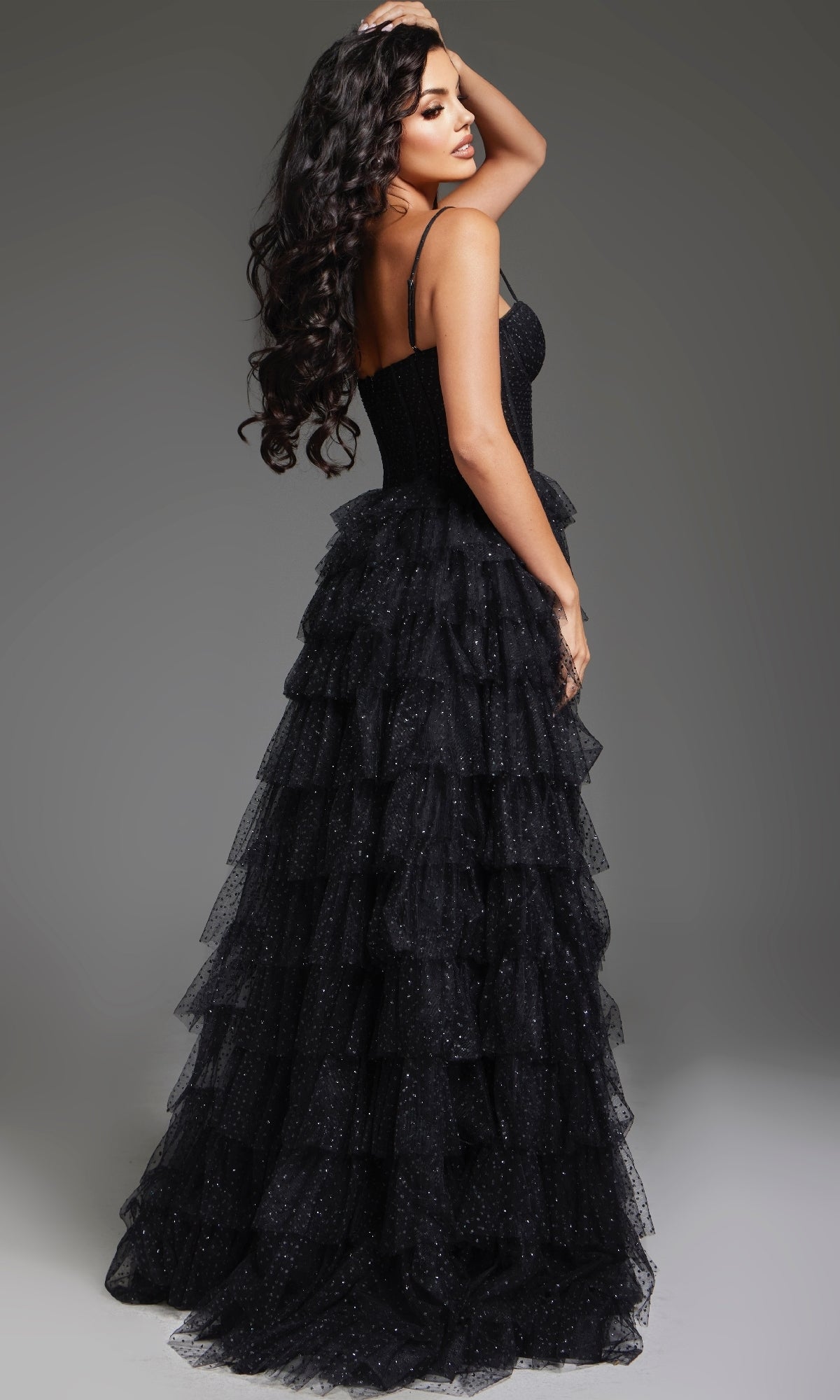 A woman in the Tiered Ruffle Black Prom Ball Gown: Jovani 38278 stands facing away, looking over her shoulder. The dress features black glitter tulle and a dramatic ruffle ball-gown skirt against a plain gray background.