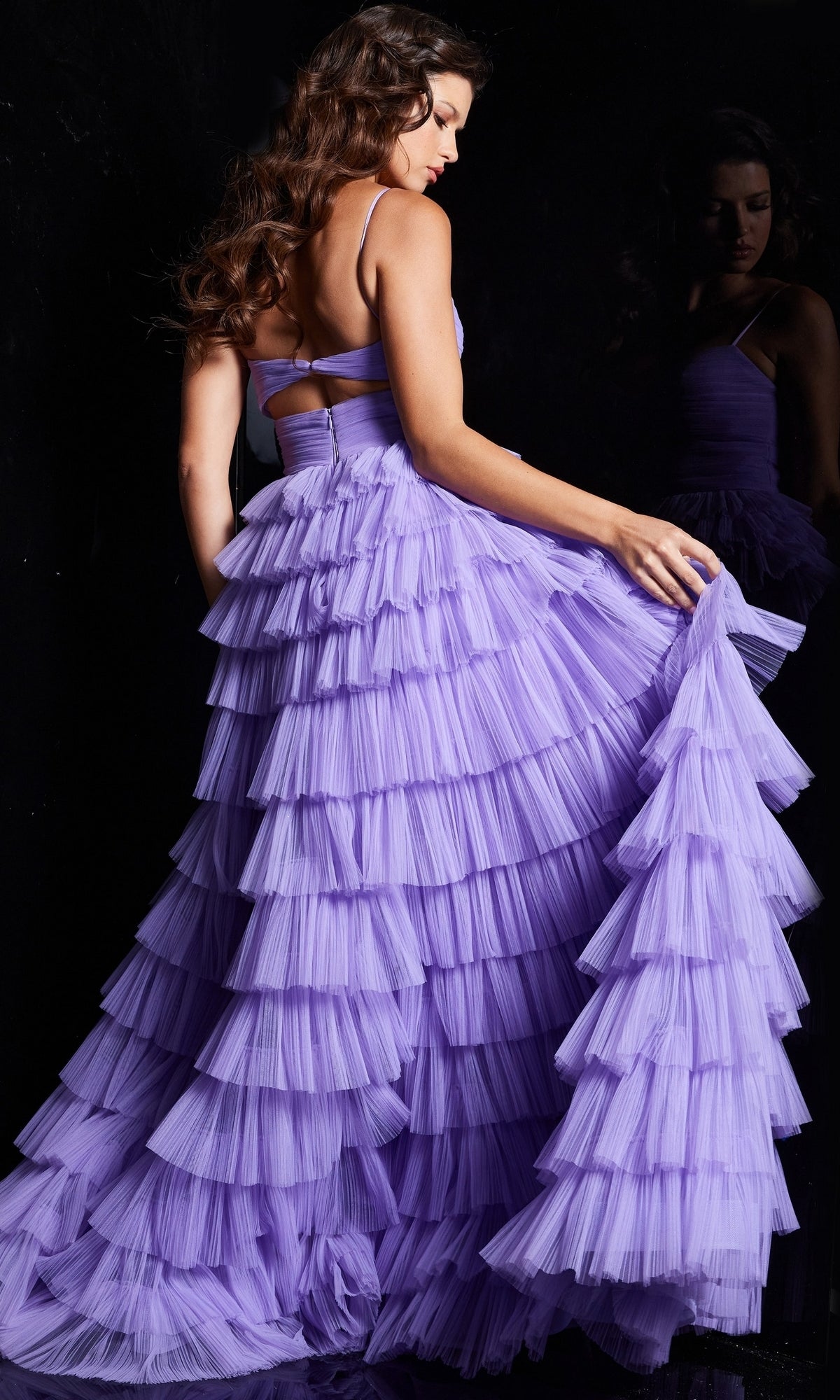 A woman in the Jovani Cut-Out Long Ruffled Prom Ball Gown 38290, featuring a tiered tulle skirt, stands with her back to the camera, holding her dress as her reflection appears in a dark mirror.