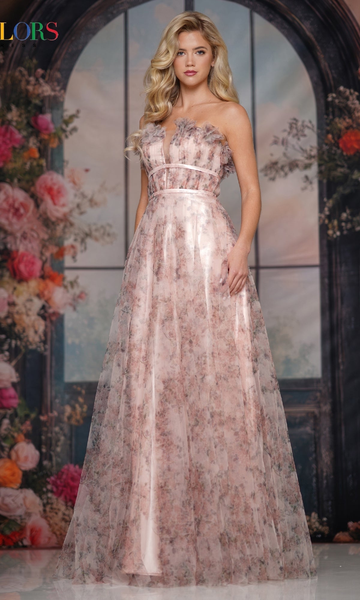 A woman poses before a floral backdrop, wearing the Colors Dress 3831—a strapless, floor-length pink chiffon prom dress with an A-line skirt and delicate 3D floral appliqués.