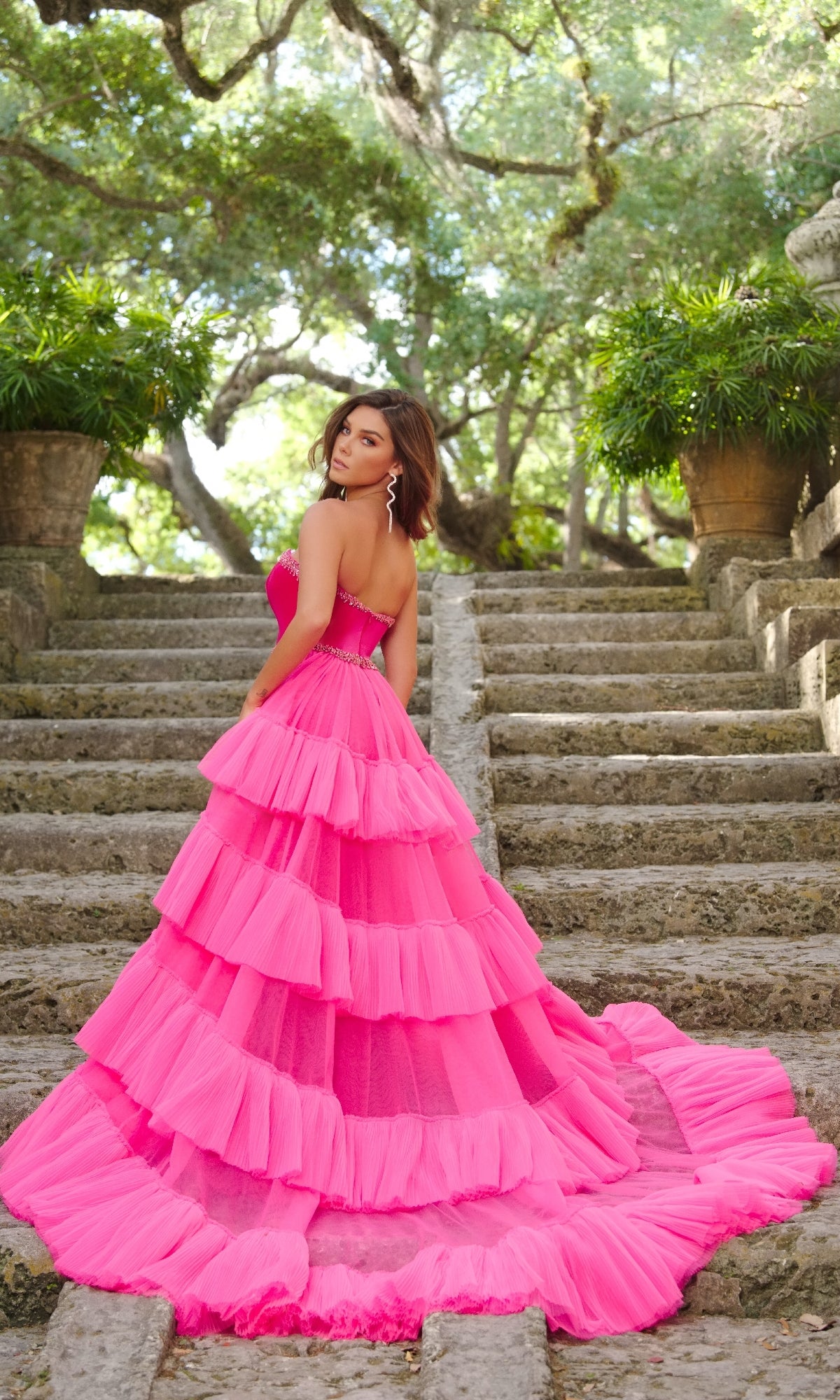 Wearing the Ava Presley 38334 strapless two-tone prom dress with a beaded bodice and tiered hot pink skirt, a woman stands on stone steps outdoors, surrounded by lush greenery and large potted plants, looking over her shoulder.