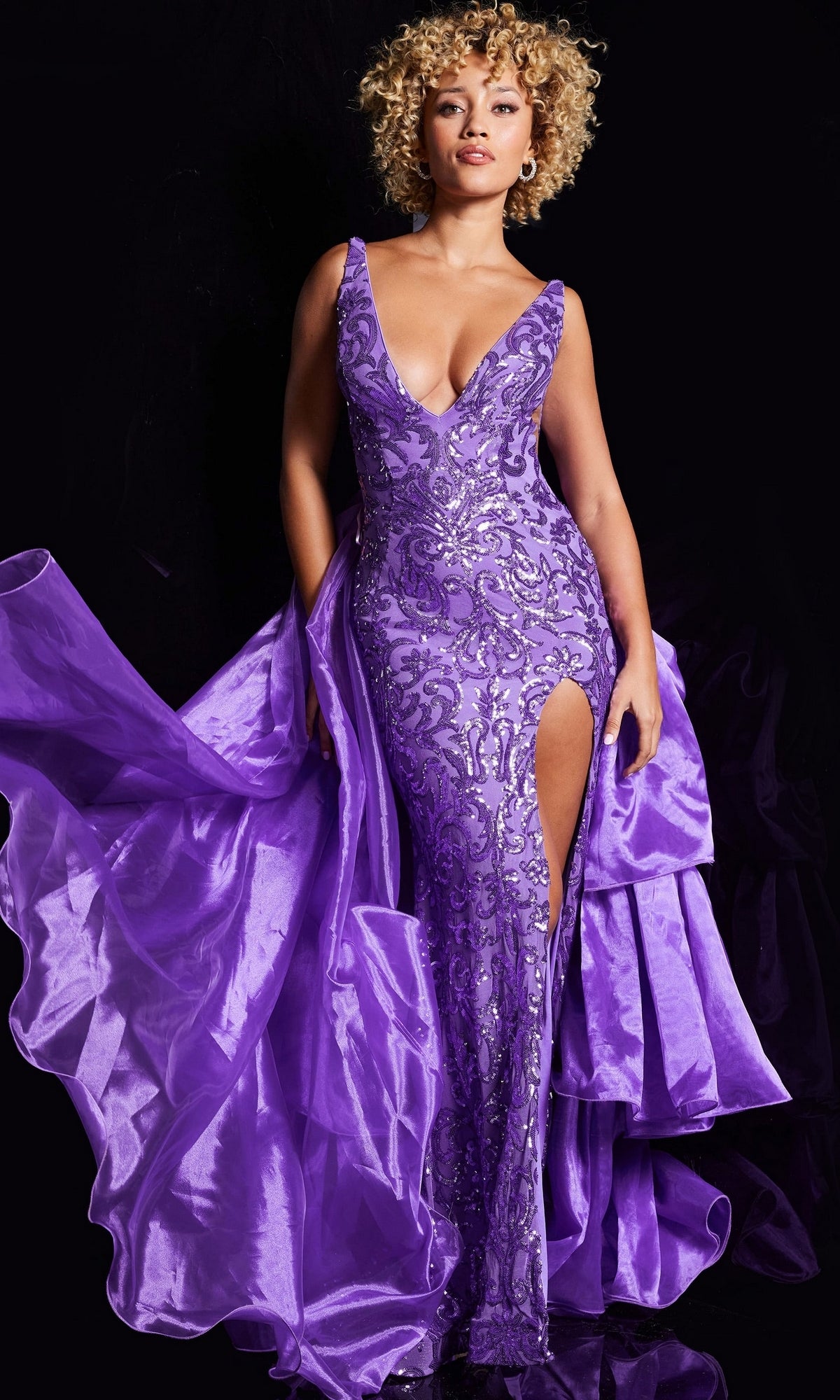 Against a black background, a person wears the Jovani Ruffled Long Sequin-Print Prom Dress 38336—a sleeveless formal gown with a deep v-neck, high slit, purple sequin print, and flowing ruffled fabric.