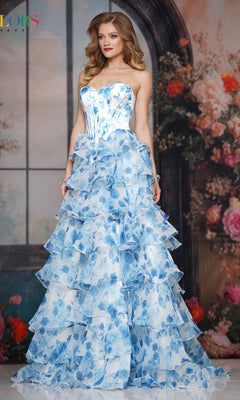 A woman stands before a floral arched window, wearing the Colors Dress 3836—a strapless, floor-length ruffle prom gown by Colors Dress. The blue and white floral pattern and her long wavy hair create an elegant, confident look.