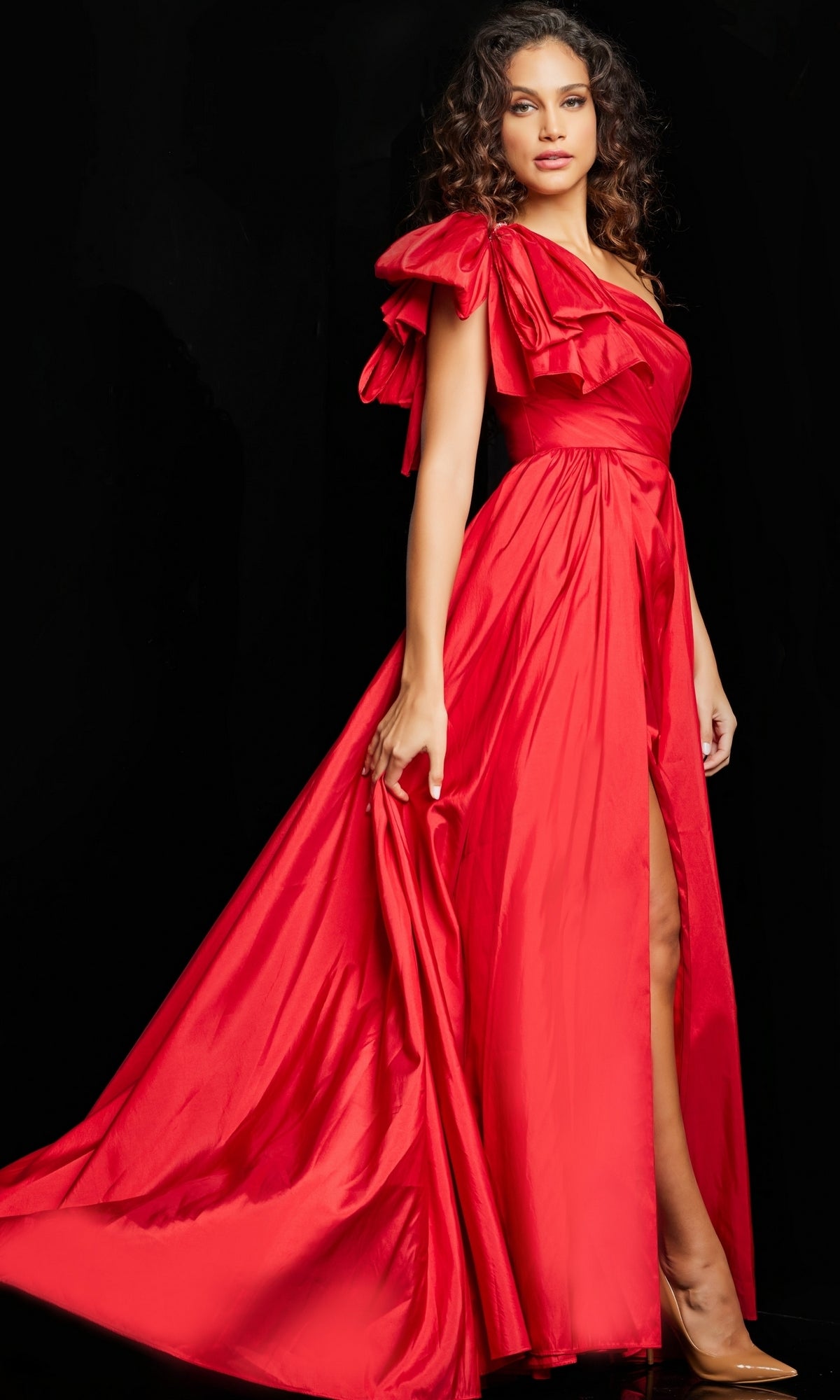 A woman poses against a black background in the Jovani One-Shoulder Bow Long Prom Dress 38466, featuring a flowing silhouette, ruffled sleeves, and a thigh-high slit.