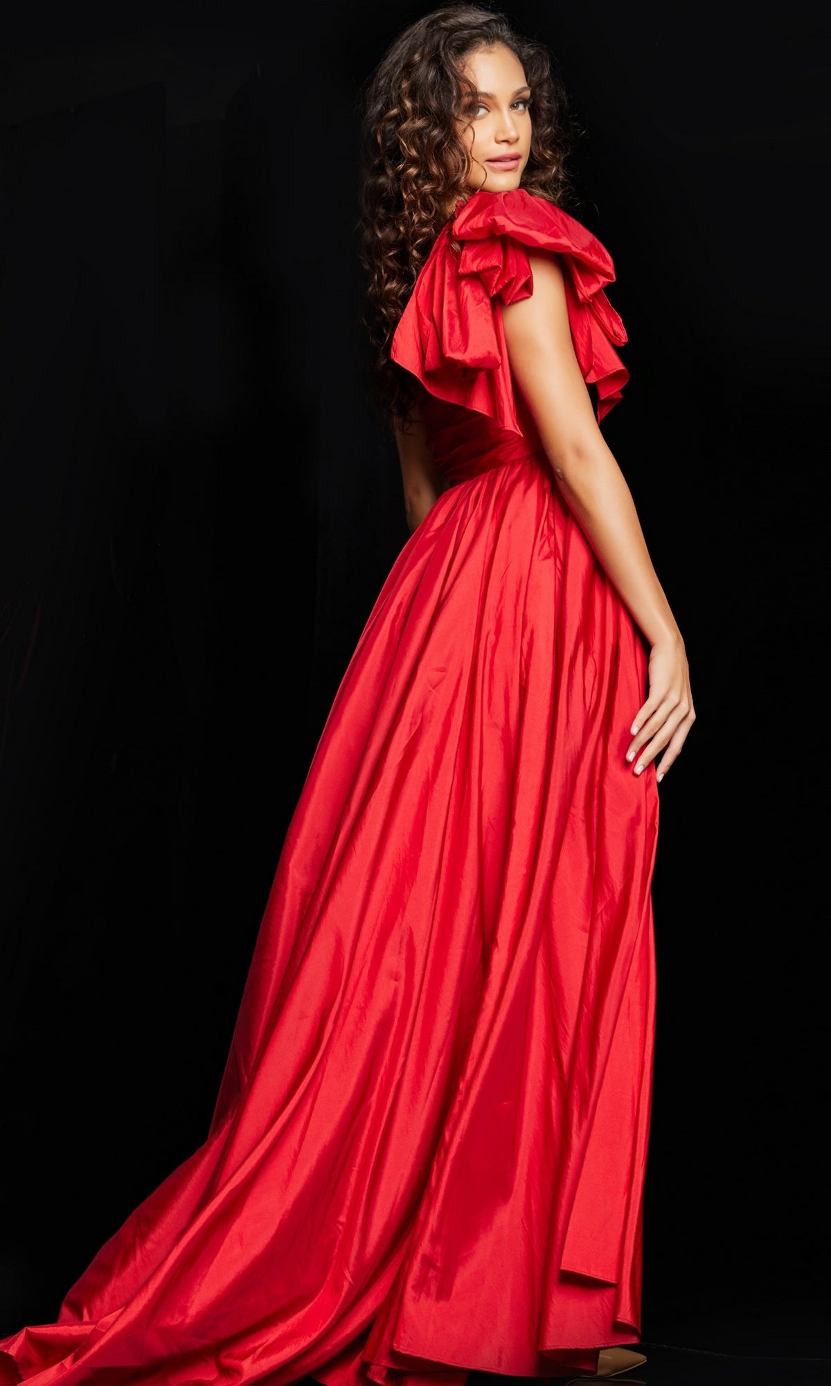 A woman with long curly hair models the Jovani One-Shoulder Bow Long Prom Dress 38466, an elegant floor-length designer gown, as she stands against a black background looking over her shoulder.