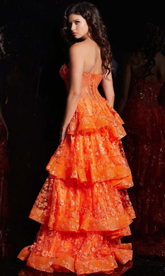 A woman with long, wavy hair wears the Jovani Strapless Long Ruffled Prom Dress 38528 in orange with floral embroidery, standing against a dark background.