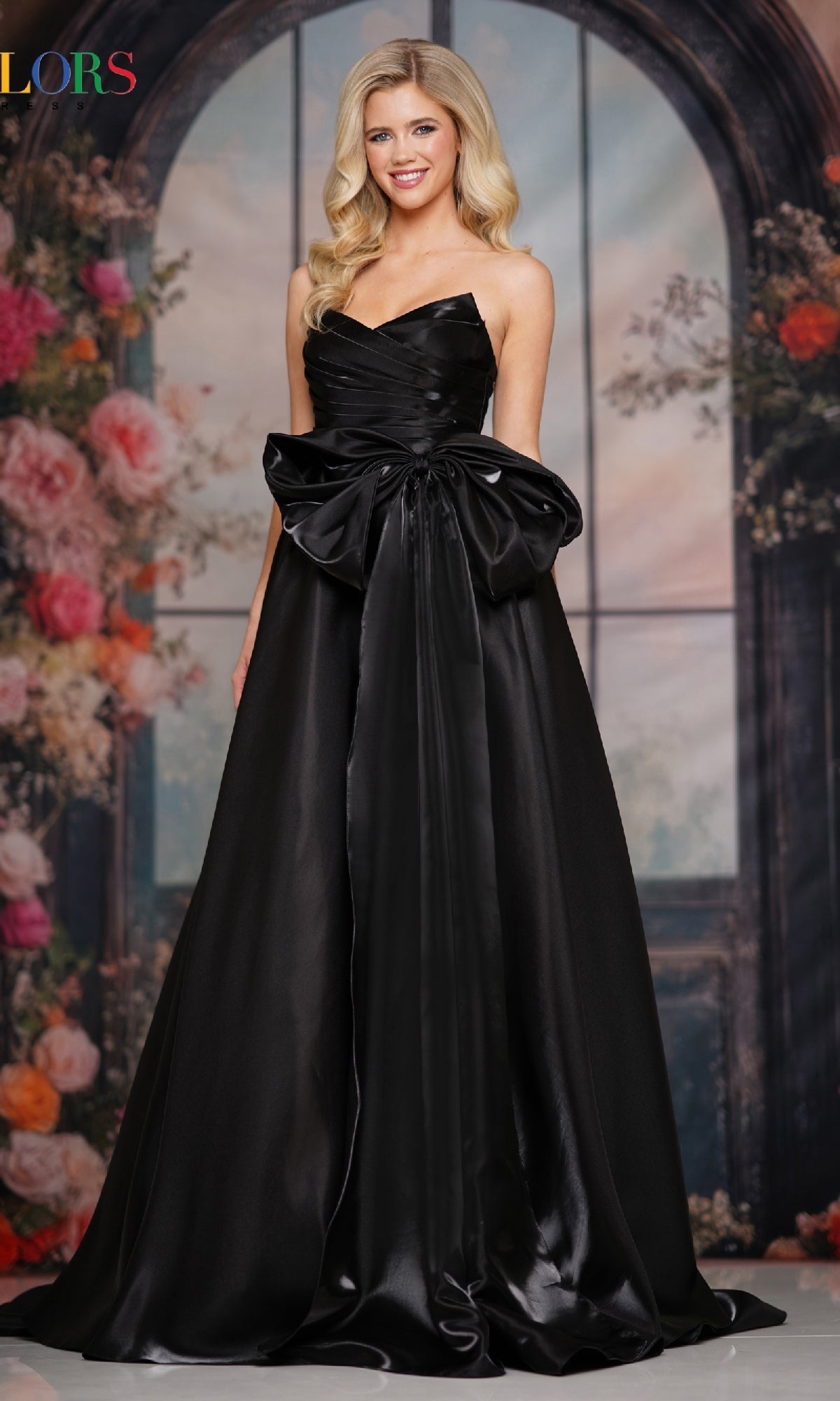A woman wearing the Lace-Up Strapless Prom Ball Gown: Colors Dress 3860 in black, featuring a corset-style bodice and large waist bow, stands before a floral and arched window backdrop.