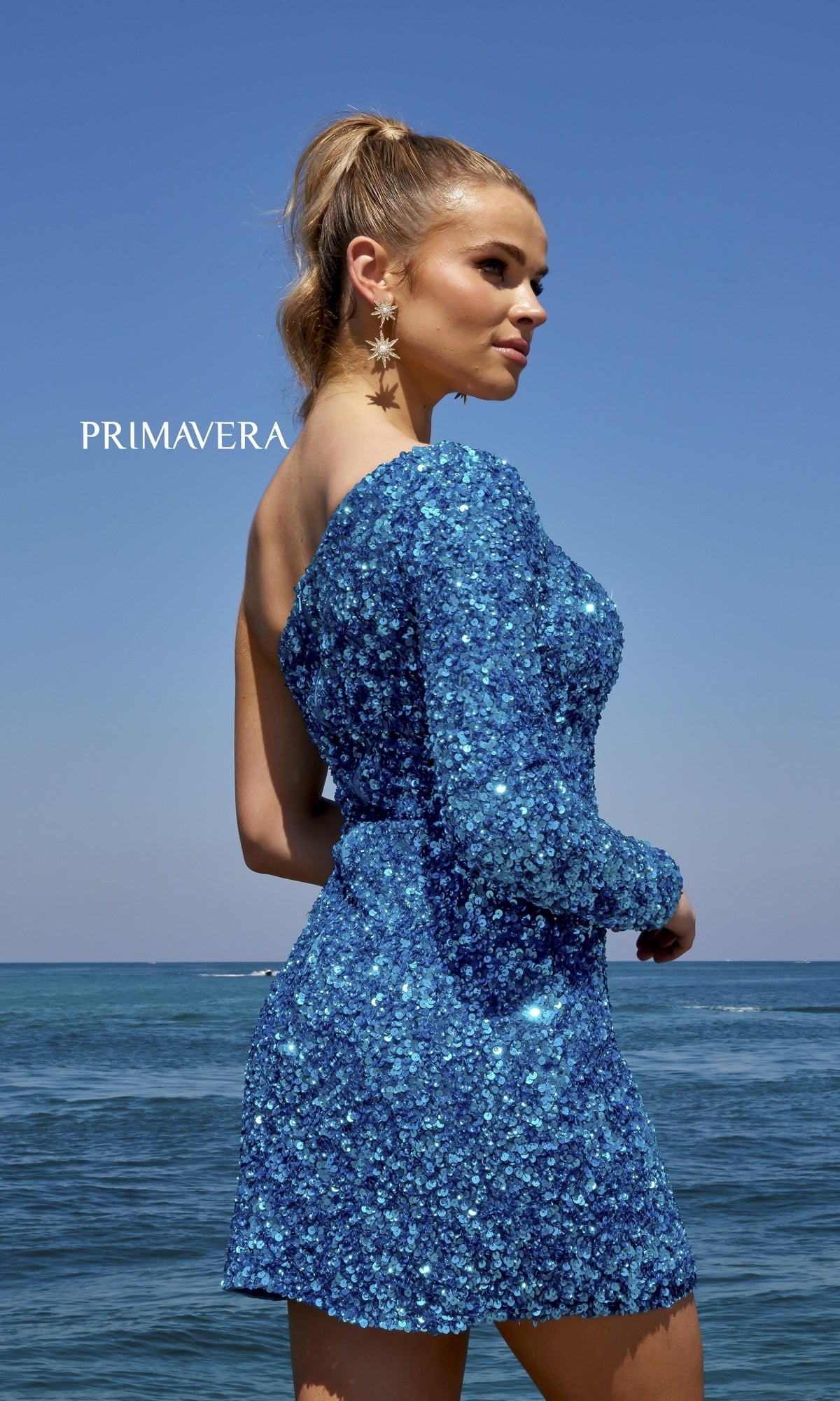 A woman in the Faux-Wrap Beaded One-Sleeve Mini Party Dress 3860 with an asymmetrical neckline stands by the ocean under a clear sky, wearing star-shaped earrings. 