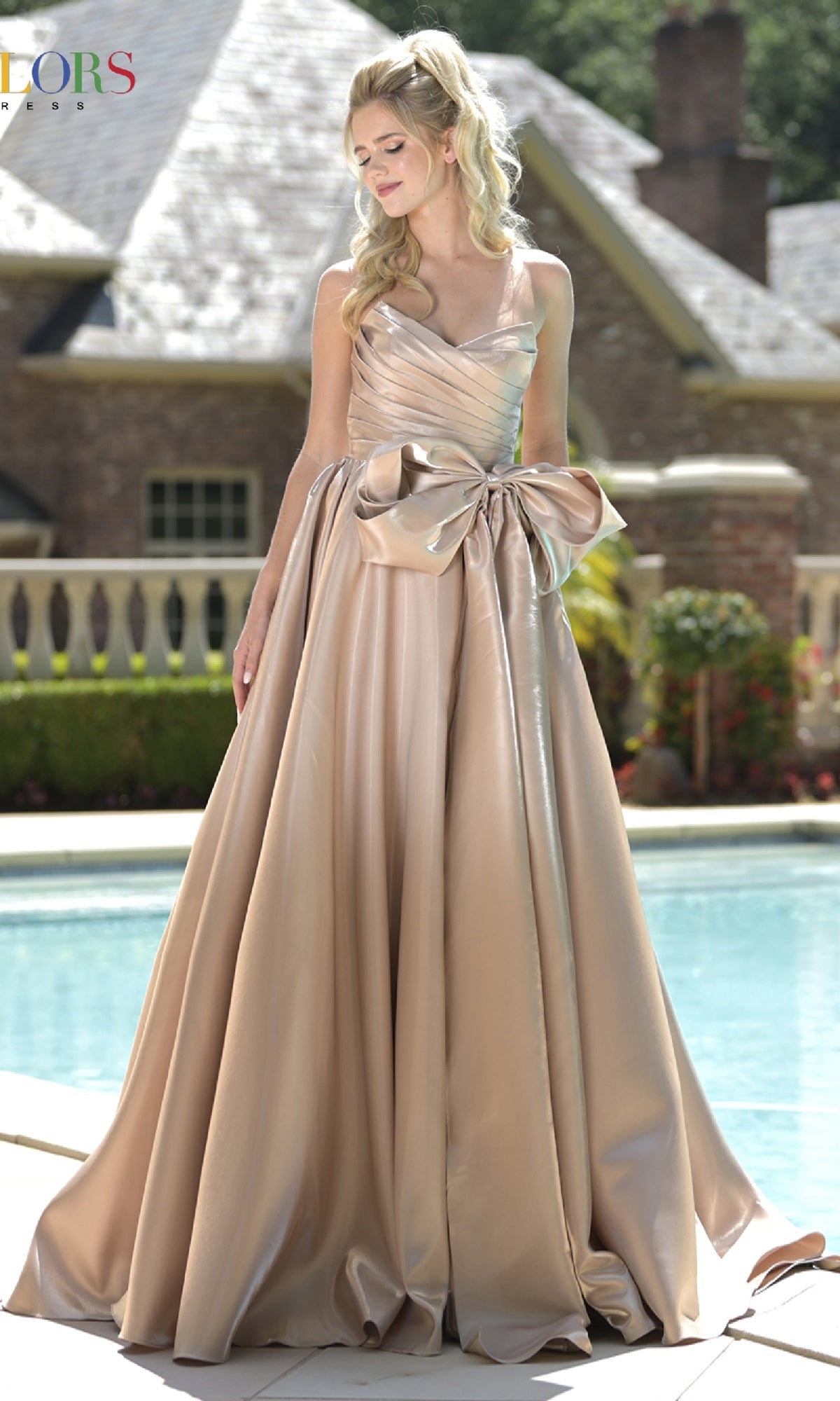 A woman in the Lace-Up Strapless Prom Ball Gown: Colors Dress 3860, a shiny champagne gown with a corset bodice and large bow, stands poolside with a brick house and garden in the background.