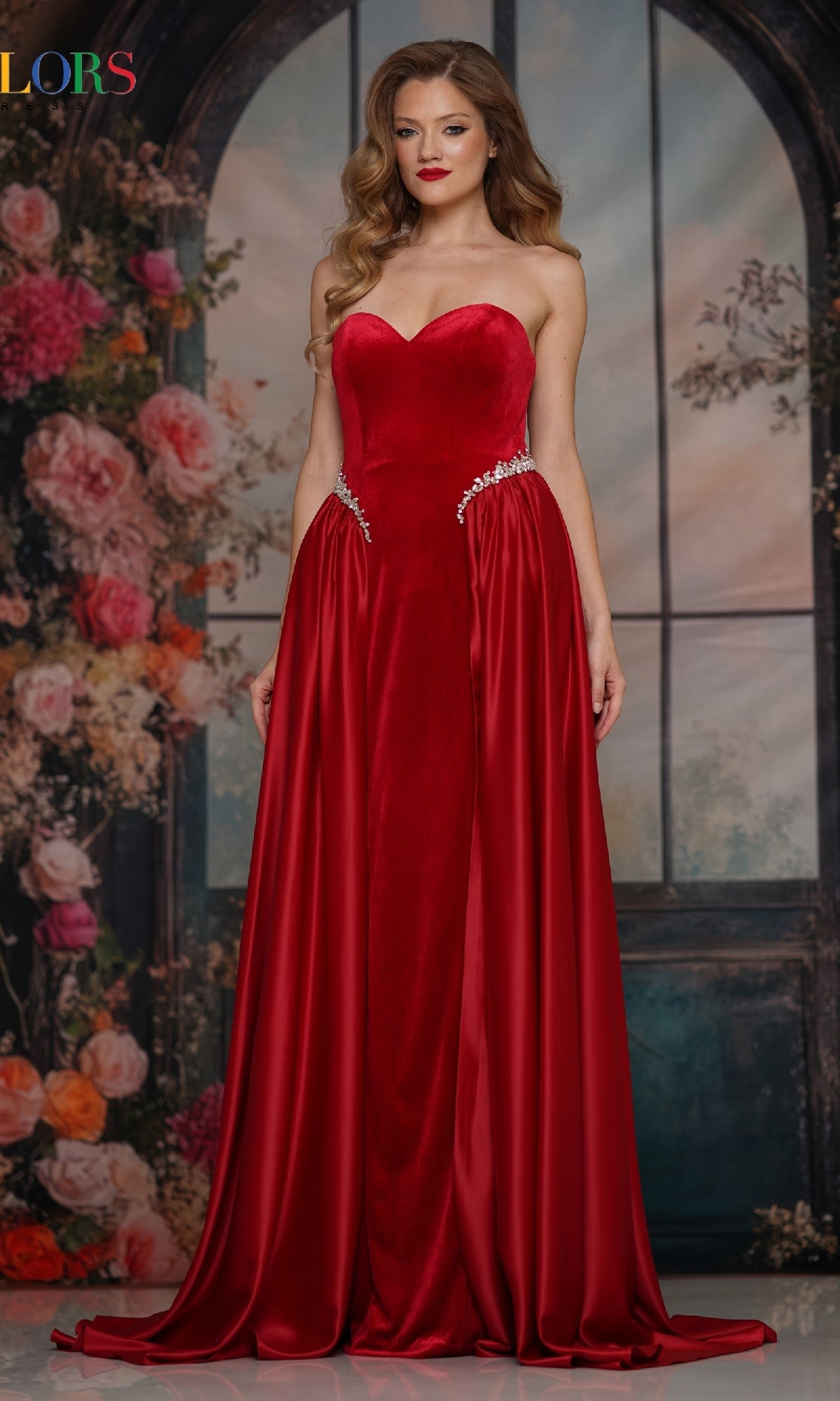 A woman in a red Strapless Velvet Prom Gown (Colors Dress 3861) featuring silver waist embellishments stands before a floral and arched window backdrop.