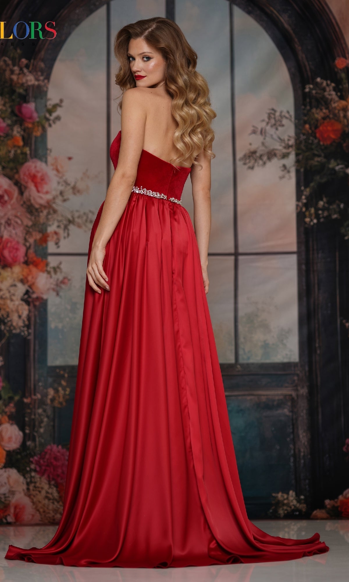 A woman models the Strapless Velvet Prom Gown: Colors Dress 3861, posing with her back slightly turned before a floral and window-themed backdrop.