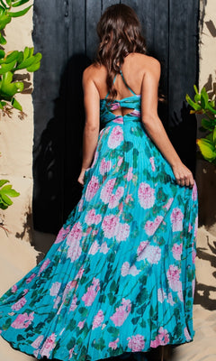 A woman stands with her back to the camera, wearing the Jovani Pleated Long Floral-Print Prom Dress 38638 in blue with pink florals, halter-neck style, holding out the skirt. Green plants frame the elegant scene.
