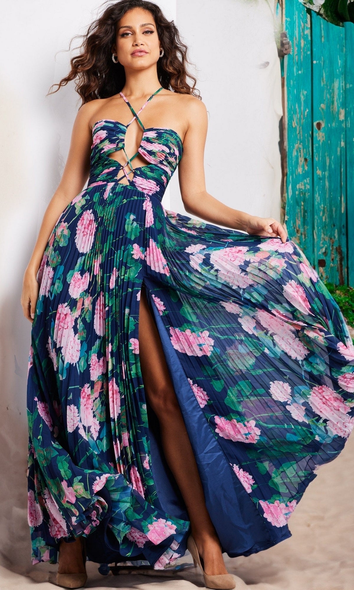 A woman stands outdoors by a turquoise door wearing the Jovani Pleated Long Floral-Print Prom Dress 38638, featuring a strapless design, pink blossom print, pleated skirt, and a high slit.