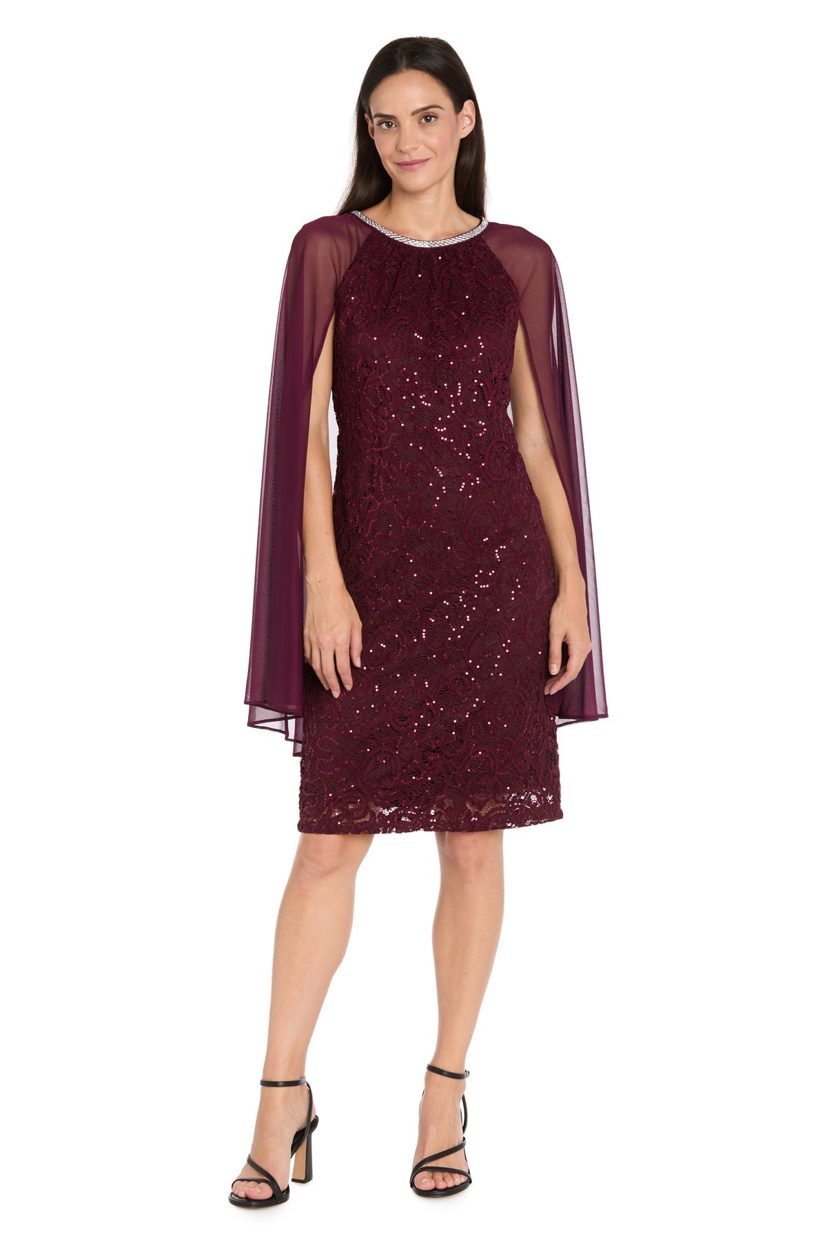 A woman stands smiling with her hands by her sides, wearing the R&M Richards 3864 Knee-Length Cape Party Dress, featuring sequined lace and a sheer cape.