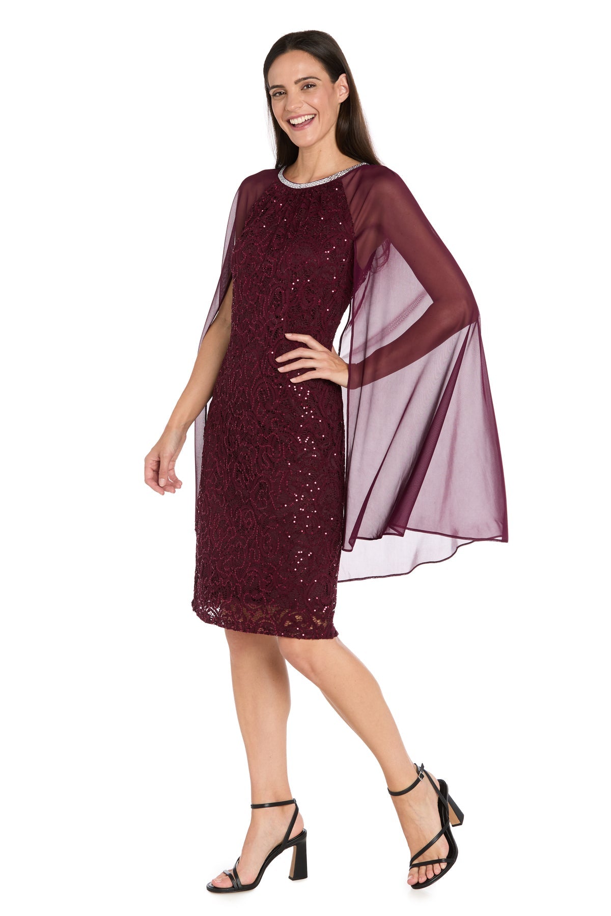 A woman in the Knee-Length Cape Party Dress: R&M Richards 3864, featuring a sequin-lace design, sheer cape overlay, and black heeled sandals, stands smiling against a white background.