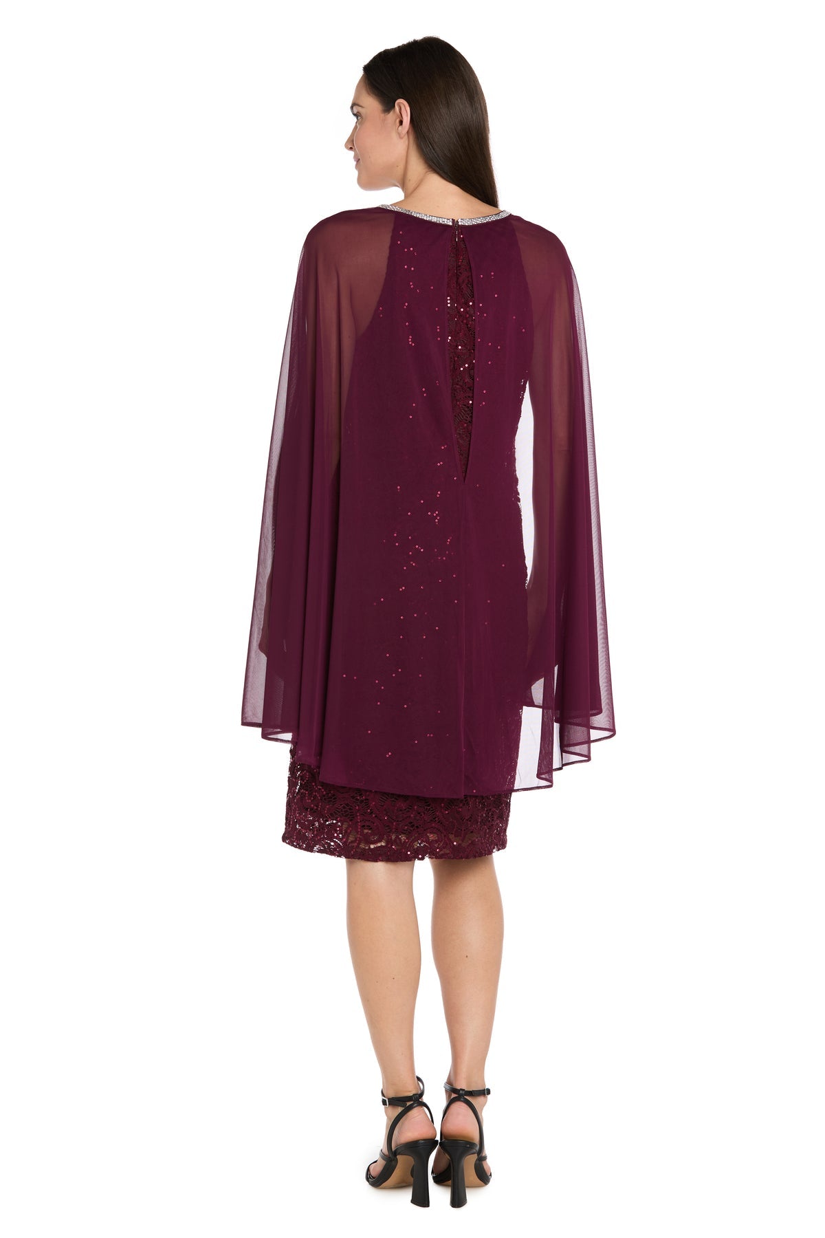 A woman faces away, wearing the R&M Richards 3864 Knee-Length Cape Party Dress with sequin lace, a sheer overlay, and black heeled sandals.