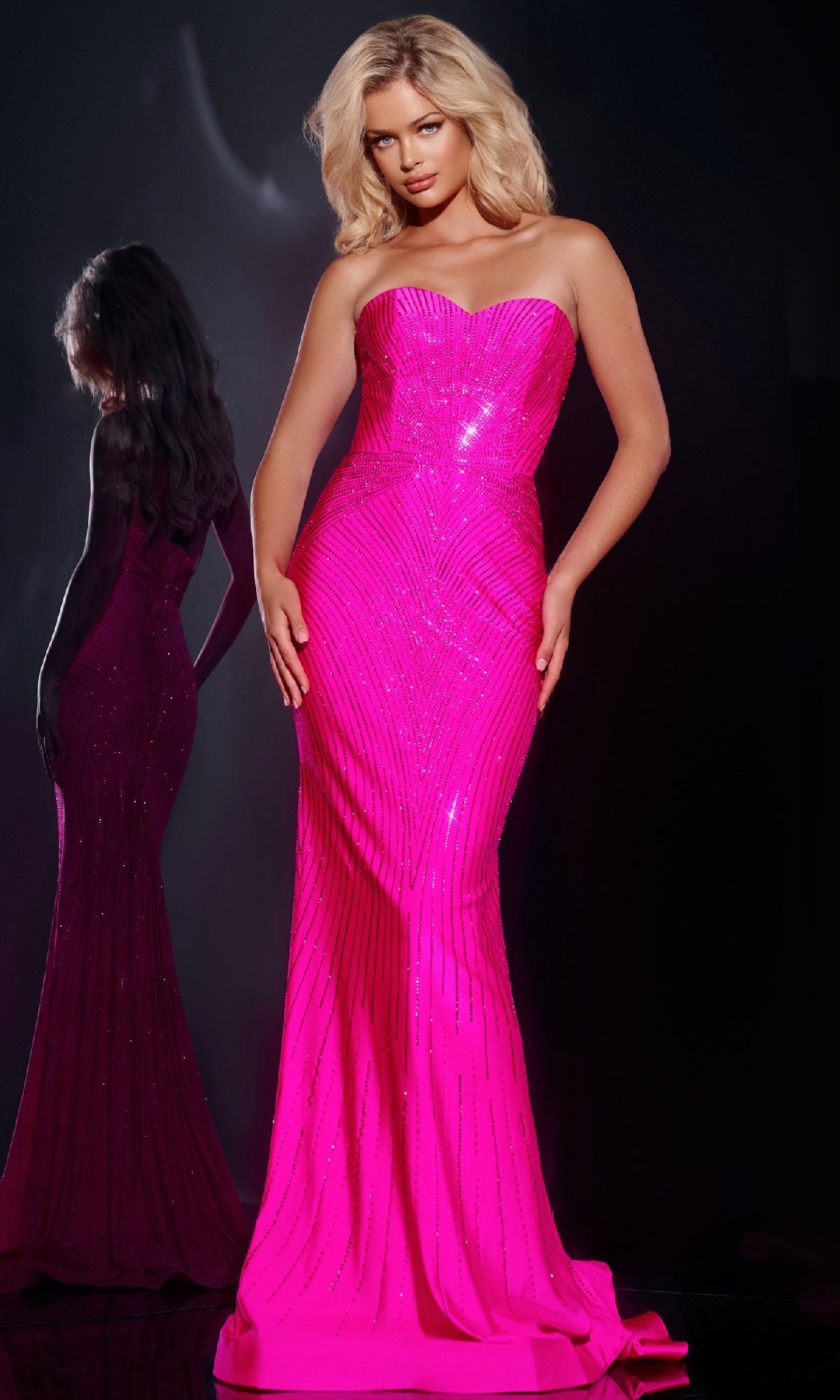 A woman stands against a dark background in the Jovani 38655 long prom dress, featuring a strapless sweetheart neckline and sparkly embellishments, with her reflection visible to the left.