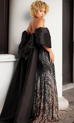 A woman with curly blonde hair wears the Caped Strapless Beaded Prom Gown (Jovani 38746), a black designer dress with an off-shoulder cape, large bow, and sequin details, standing outdoors and looking over her shoulder.