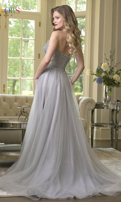 A woman with long blonde hair models the Colors Dress 3879, a strapless prom dress with an embellished corset bodice and tulle A-line skirt, standing in an elegant room with large windows, a sofa, and flowers on a side table.