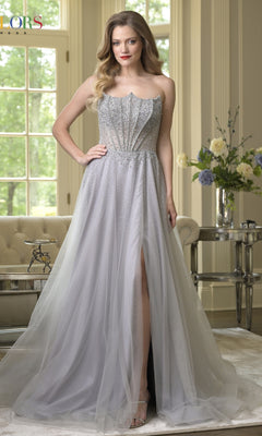 A woman stands indoors wearing the Long Prom Dress: Colors Dress 3879, featuring a strapless embellished corset bodice and a silver-gray tulle A-line skirt. The scene is set with large windows, flowers, and a mirrored table in the background.