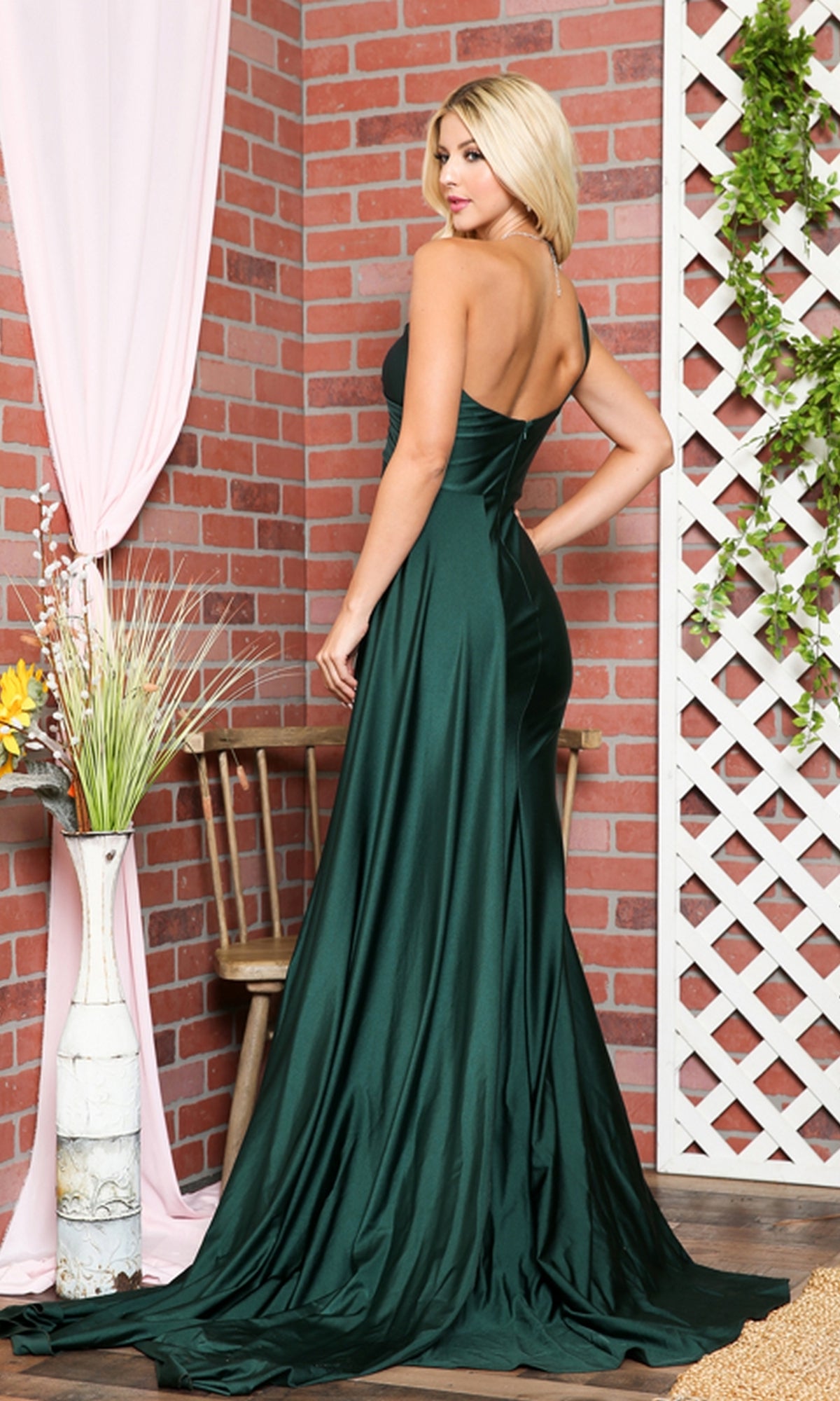 A woman in a dark green satin Side-Cape One-Shoulder Long Prom Dress 387 stands with her back to the camera against a brick wall and wooden lattice backdrop.